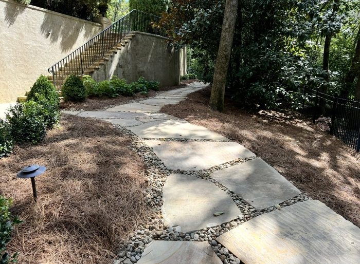 Shade garden with pine straw beds and river rock edging along a stone walkway beneath mature trees