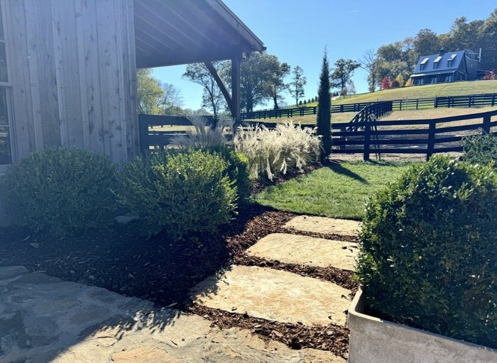 Boxwoods and ornamental grass framing stone steps beside a barn, with fenced pasture beyond